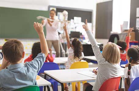 kids raising hands for teacher next to chalkboard